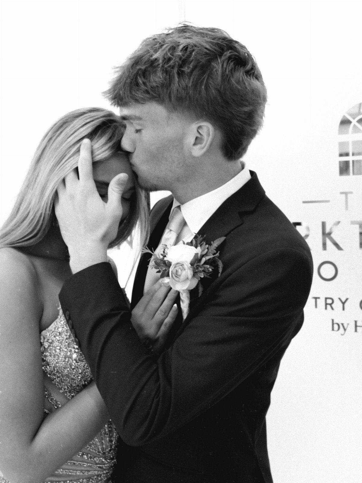 Young couple in formal wear with man kissing woman's forehead in black and white.