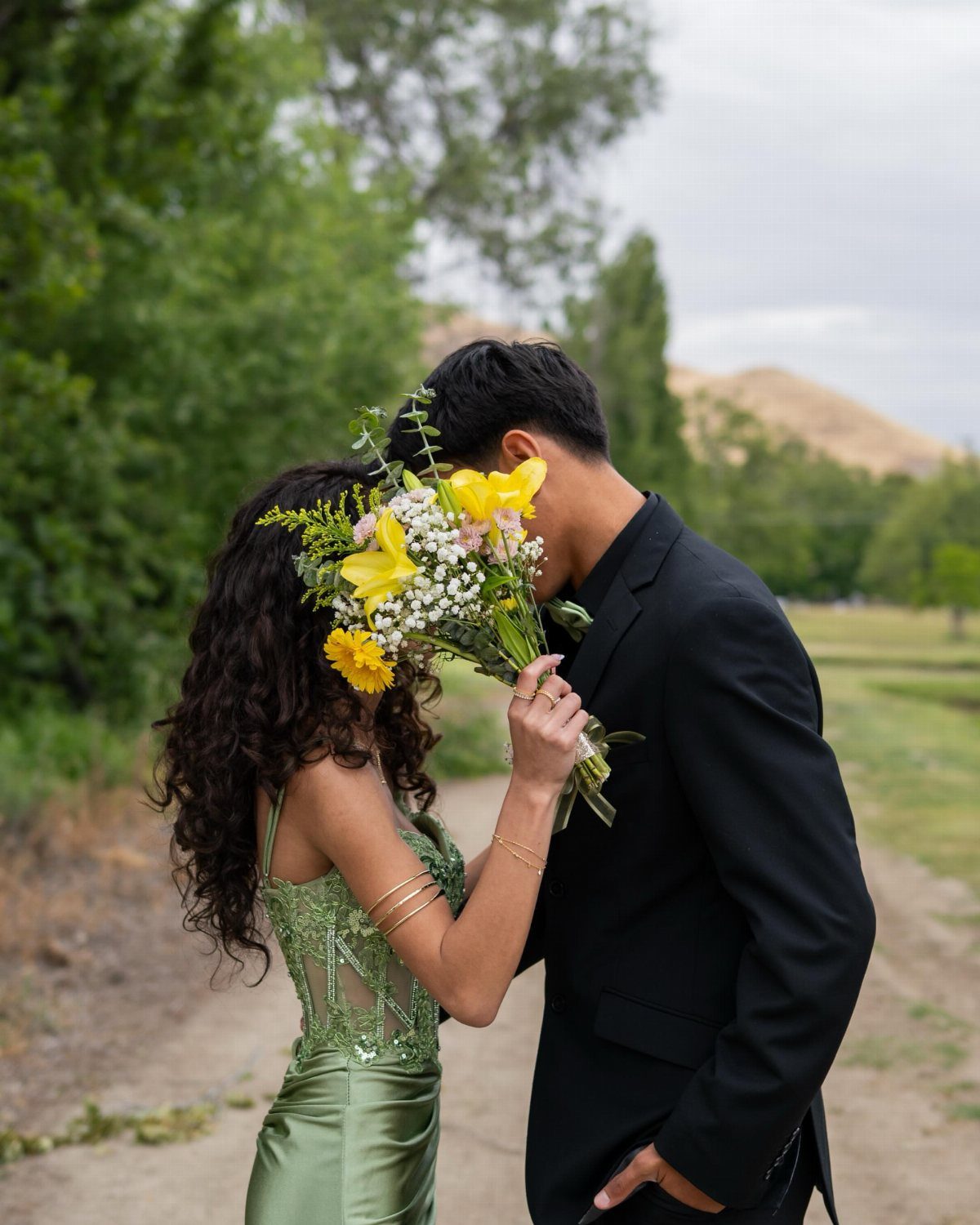 Couple in formal wear hiding their faces behind a yellow flower bouquet outdoors.