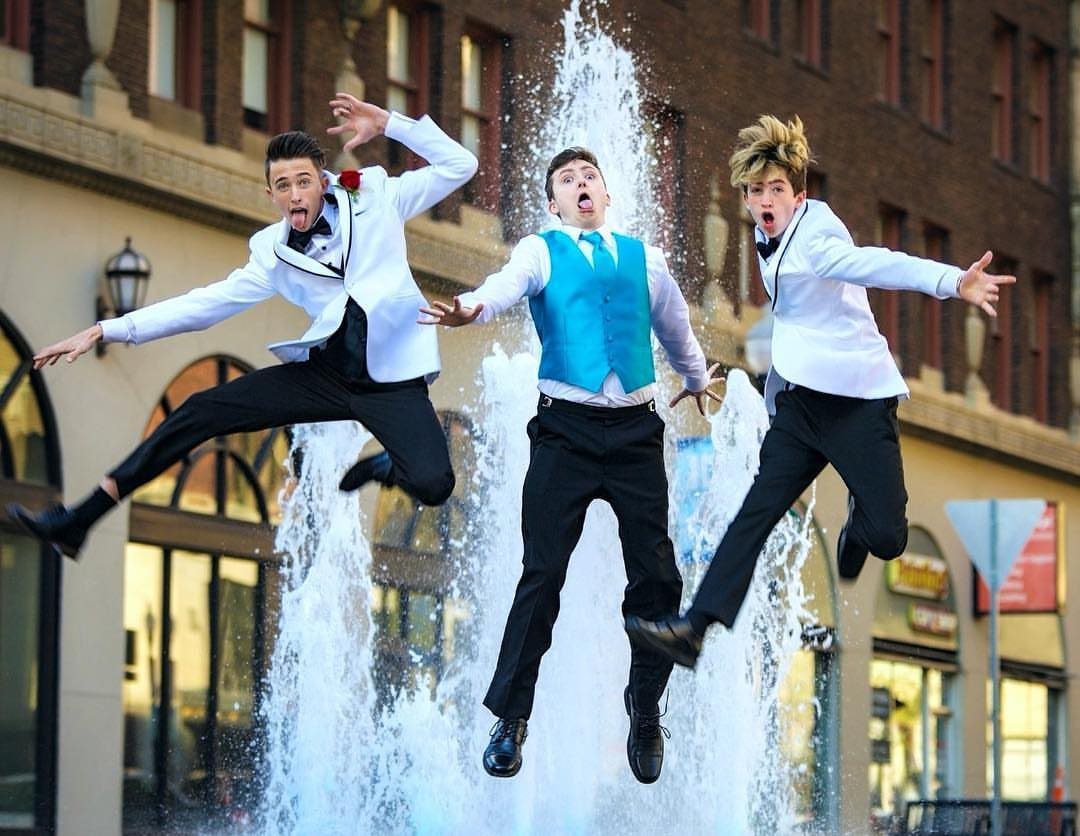 Three young men in formal wear jumping excitedly in front of a water fountain.