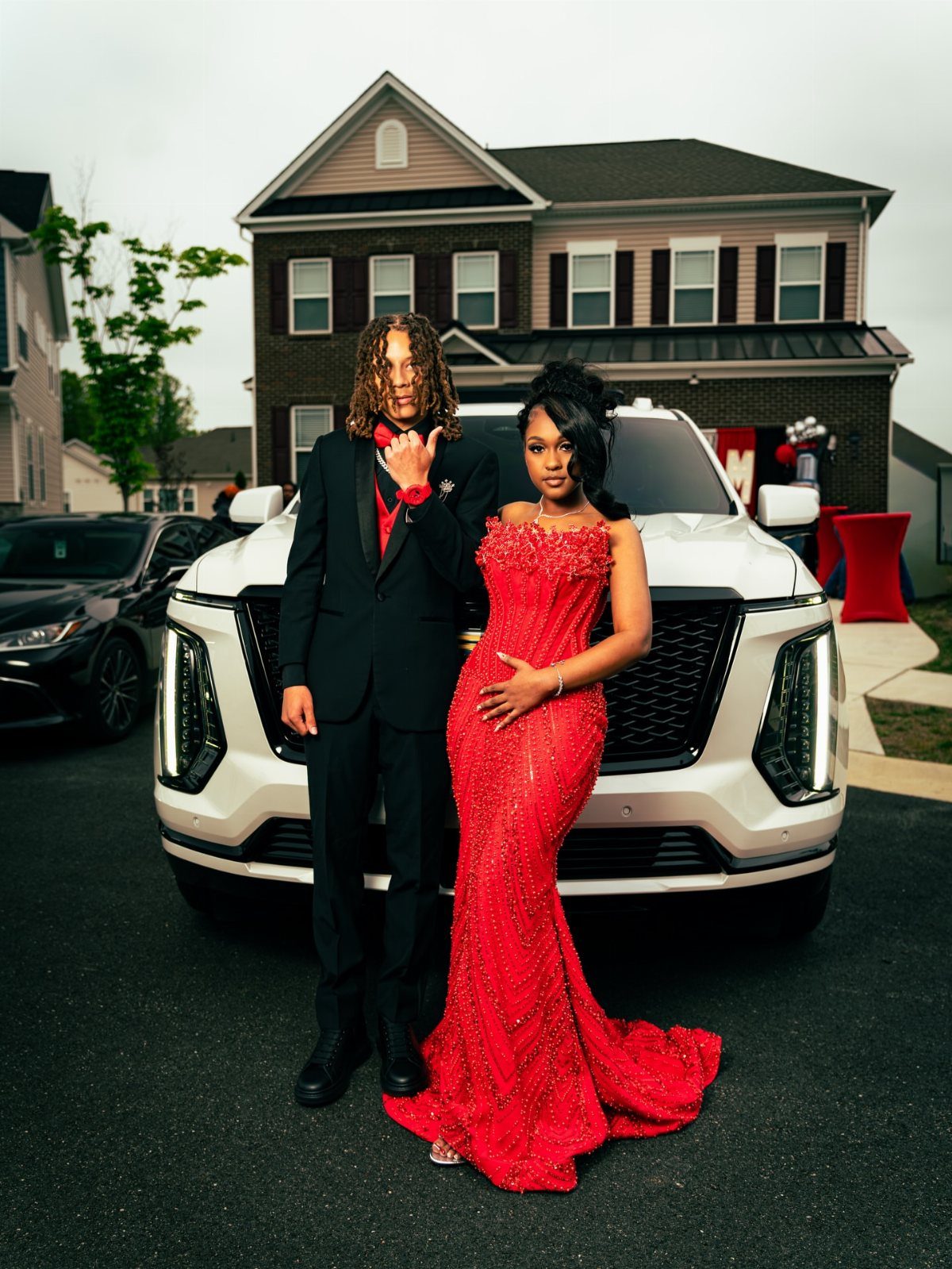 Couple in formal red and black attire posing before a white luxury SUV.