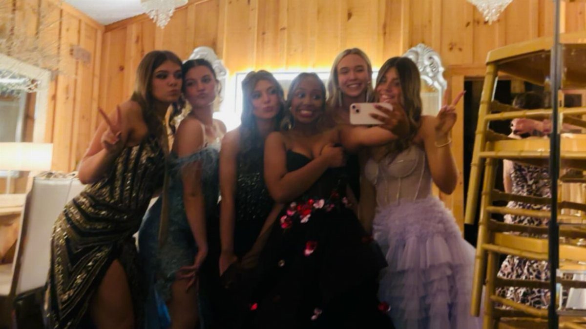 Six young women in formal dresses posing for a group selfie in a room.