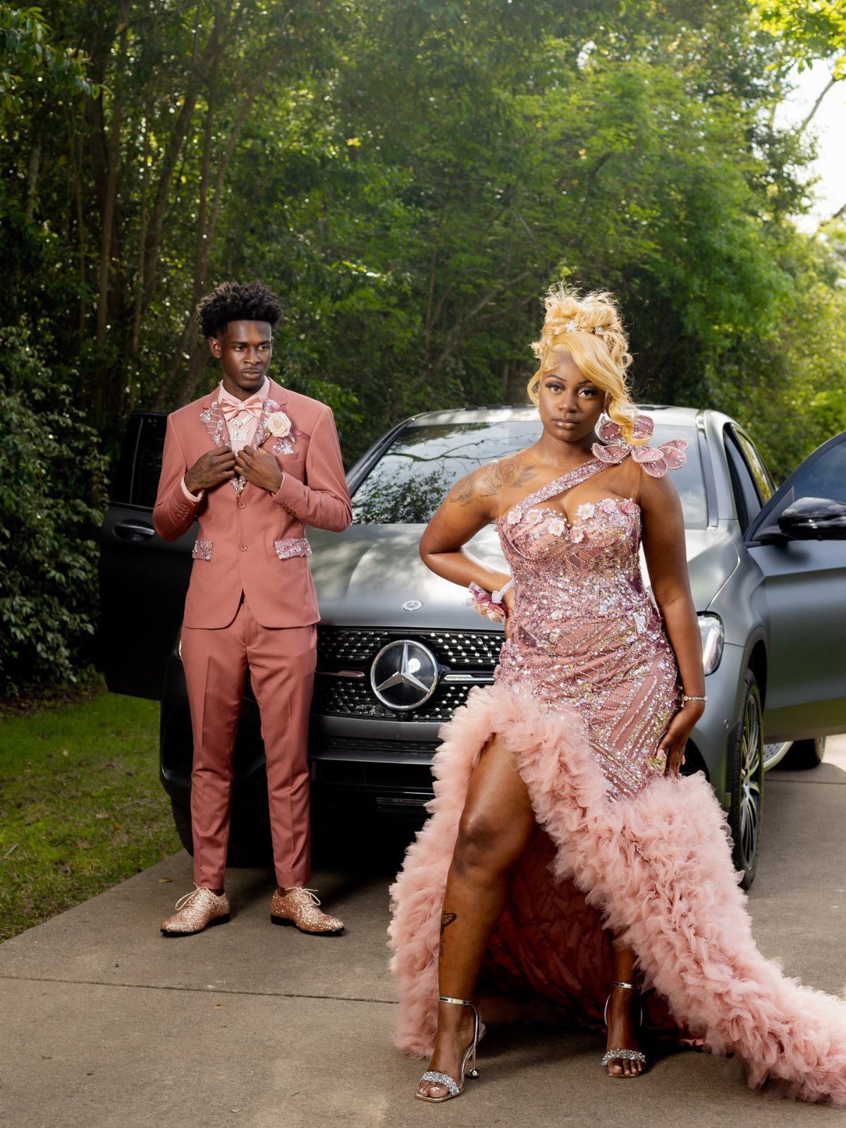 Couple in pink formal wear posing in a V-shape before a grey car.
