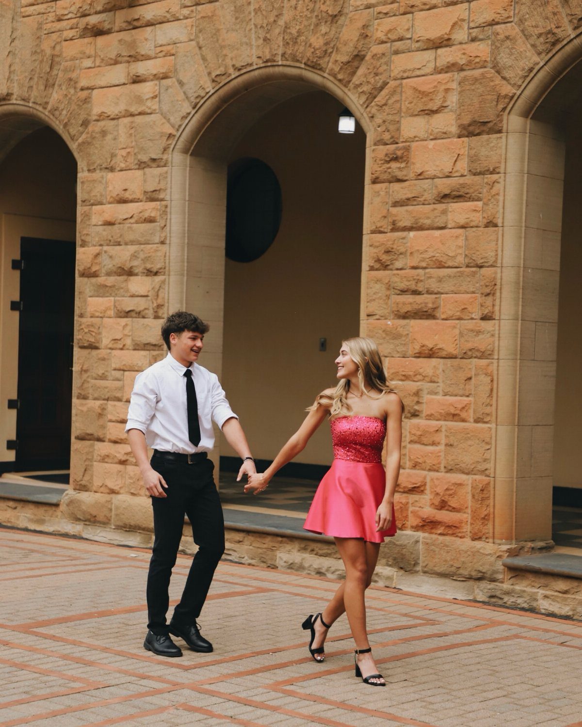 Young couple in formal attire holding hands while walking toward the camera outdoors.