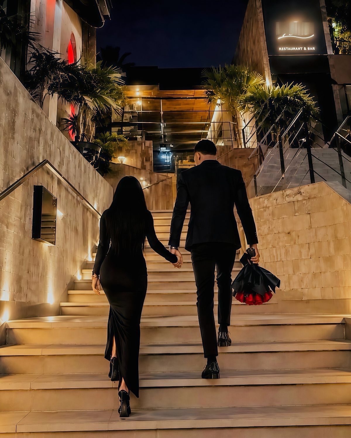 Couple in black formal wear holding hands while walking up lit stone stairs.