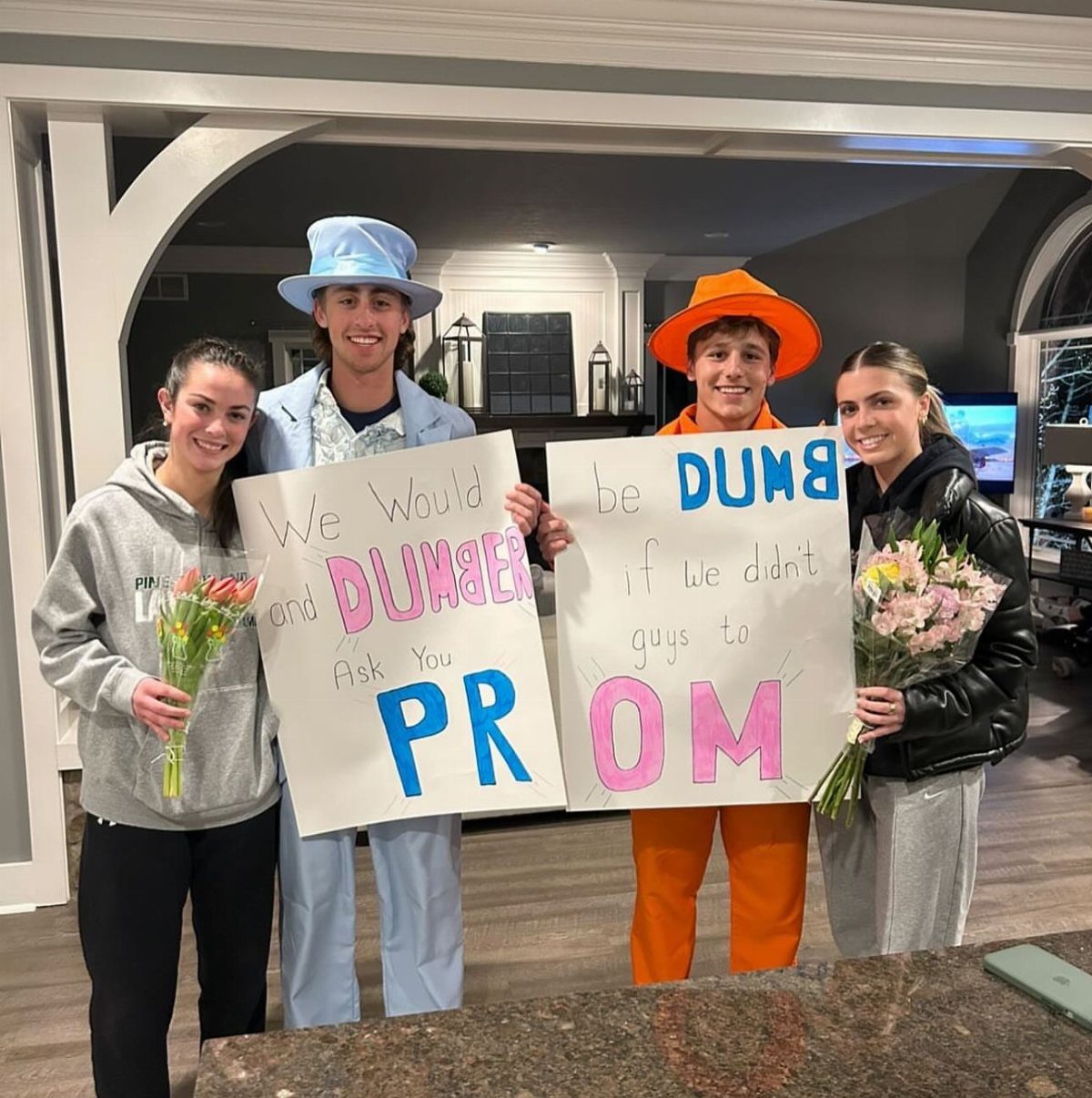 Four teenagers posing with a Dumb and Dumber themed prom proposal sign and flowers.