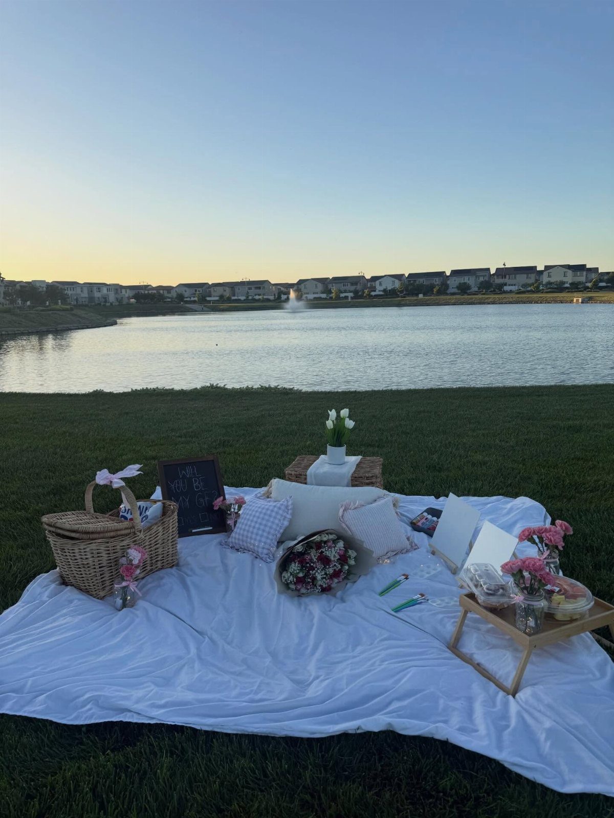 Picnic setup with white blanket, flowers, and basket by a calm lake at sunset.