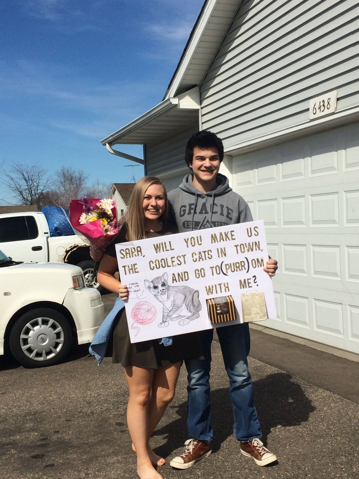 Teenage couple posing in a driveway with a cat-themed prom proposal poster.