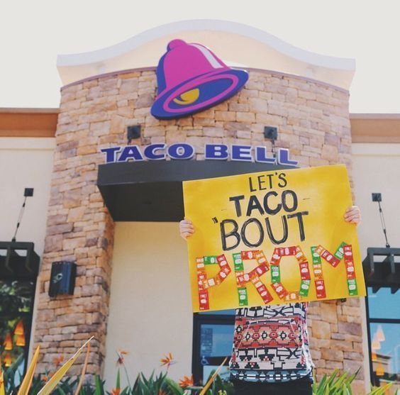Person holding a yellow taco-themed prom proposal poster in front of Taco Bell.