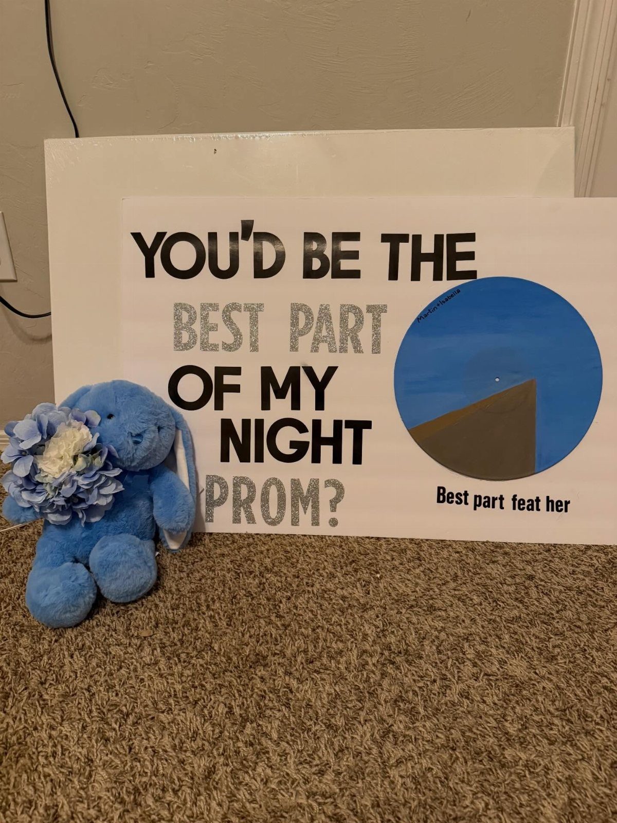Blue teddy bear with flowers sitting next to a prom proposal poster.