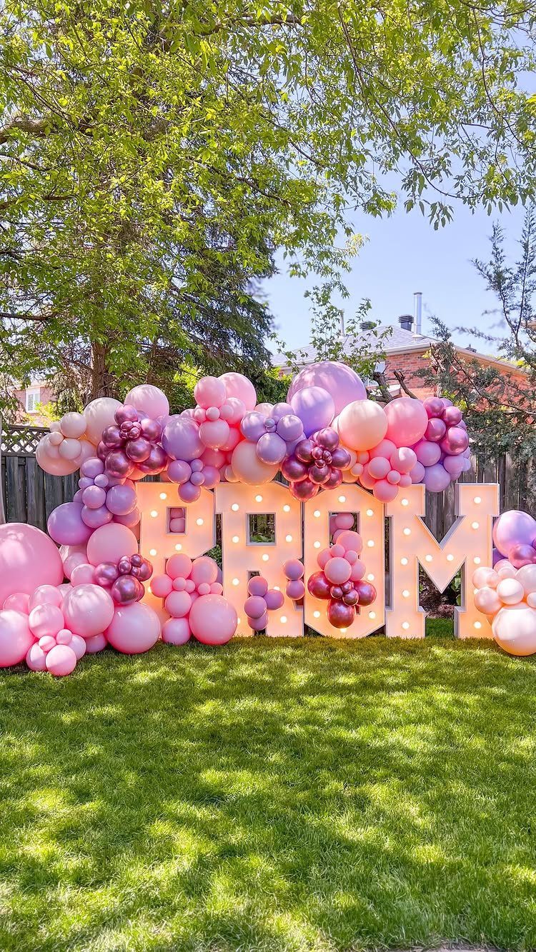 Illuminated PROM letters surrounded by pink and purple balloons on a green lawn