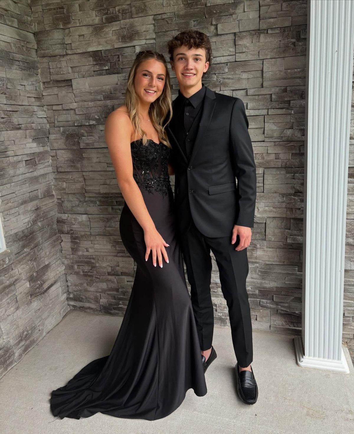 Young couple posing in formal black attire against a grey stone wall.