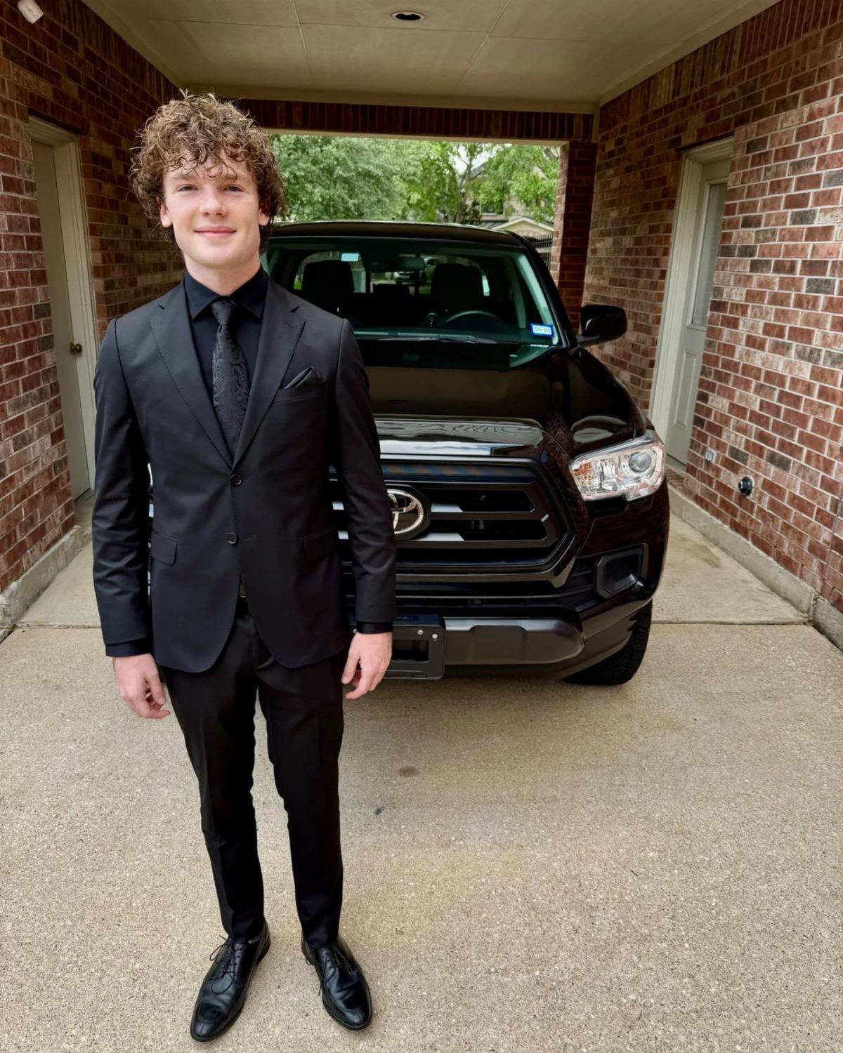 Young man in a black suit and tie standing before a black truck.