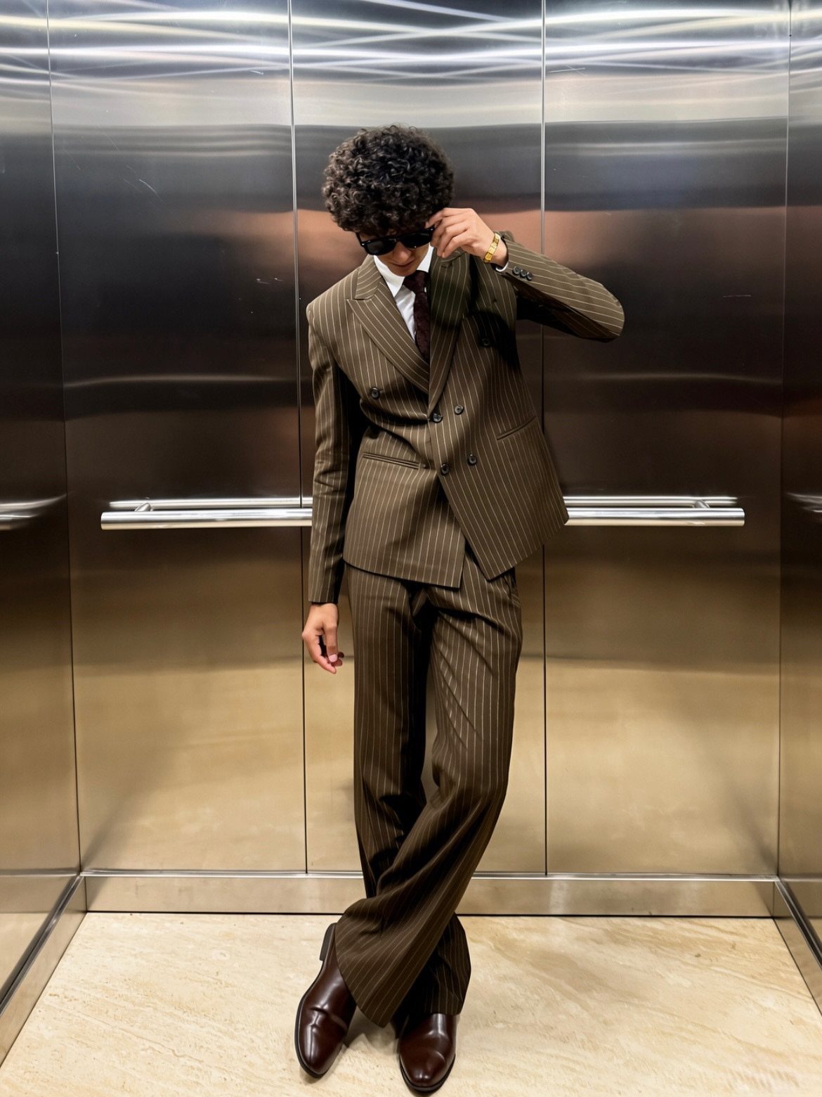 Man in a brown pinstripe suit and sunglasses posing inside a metallic elevator