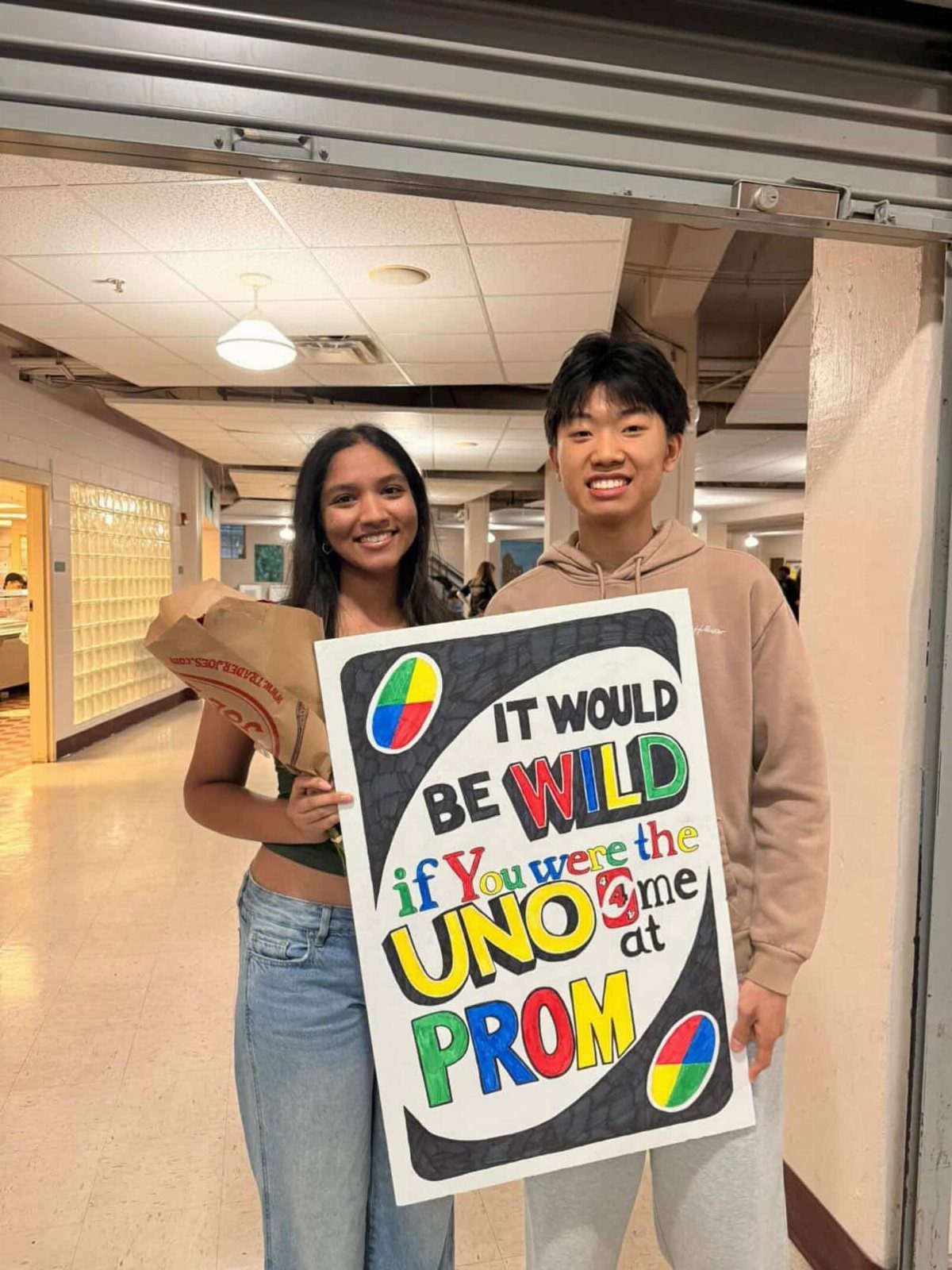 Teenage couple posing in a school hallway with a colorful promposal poster.