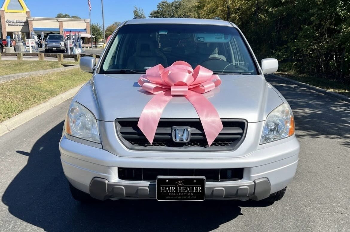 Silver car with a large pink ribbon tied on the hood outdoors