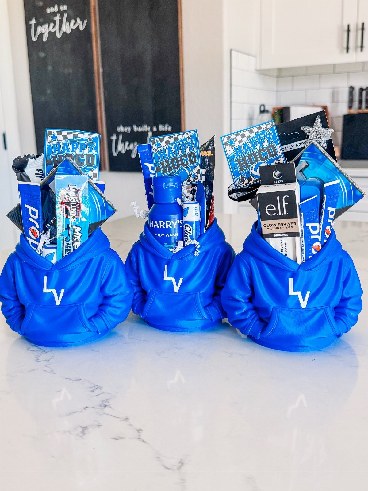 Three blue hoodies filled with blue-themed gift items on a white marble counter.