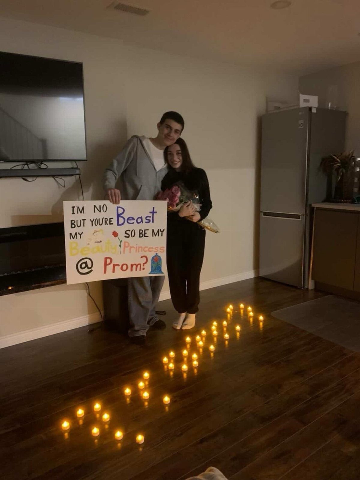 Teenage couple posing with a Beauty and the Beast promposal sign and floor candles.