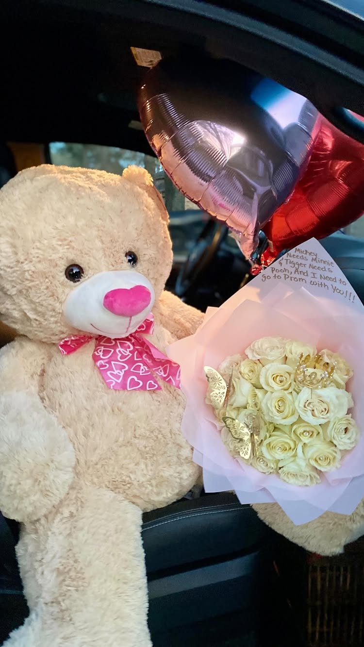 Teddy bear holding white roses and a red heart balloon inside a car