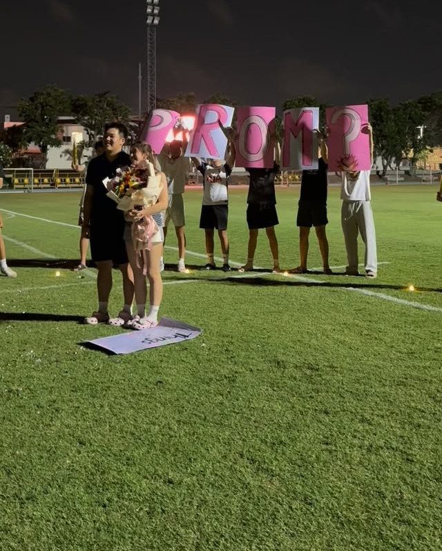 Couple on a sports field with friends holding pink letters spelling PROM?