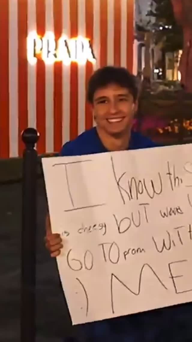 Smiling young man holding a handmade promposal sign in front of a Prada store.