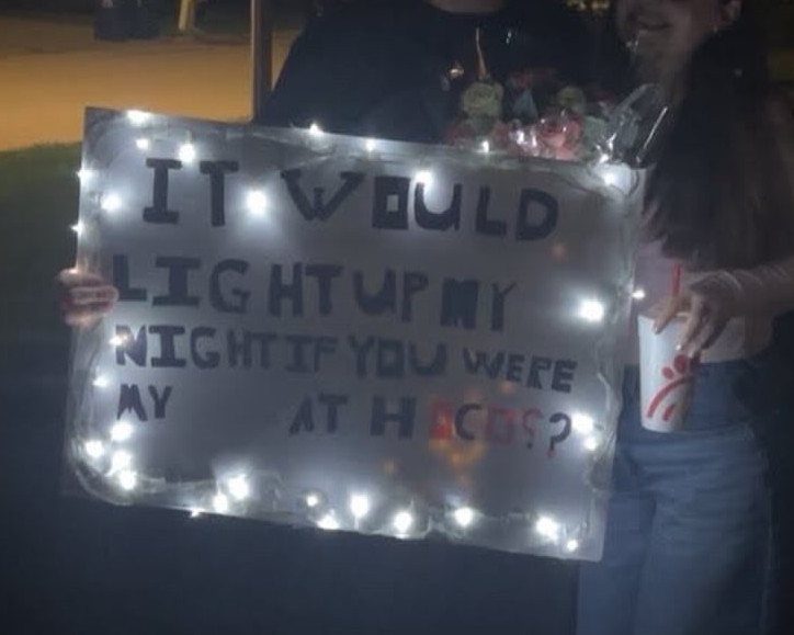 Handwritten promposal sign bordered with glowing white fairy lights held by two people.