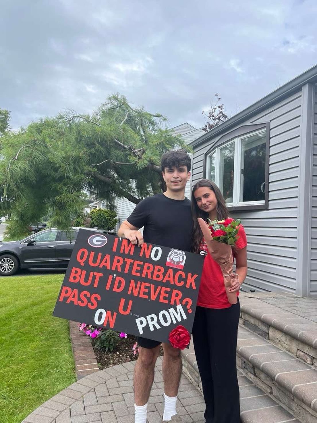 Teenage couple posing with a football themed promposal sign and red roses outdoors.