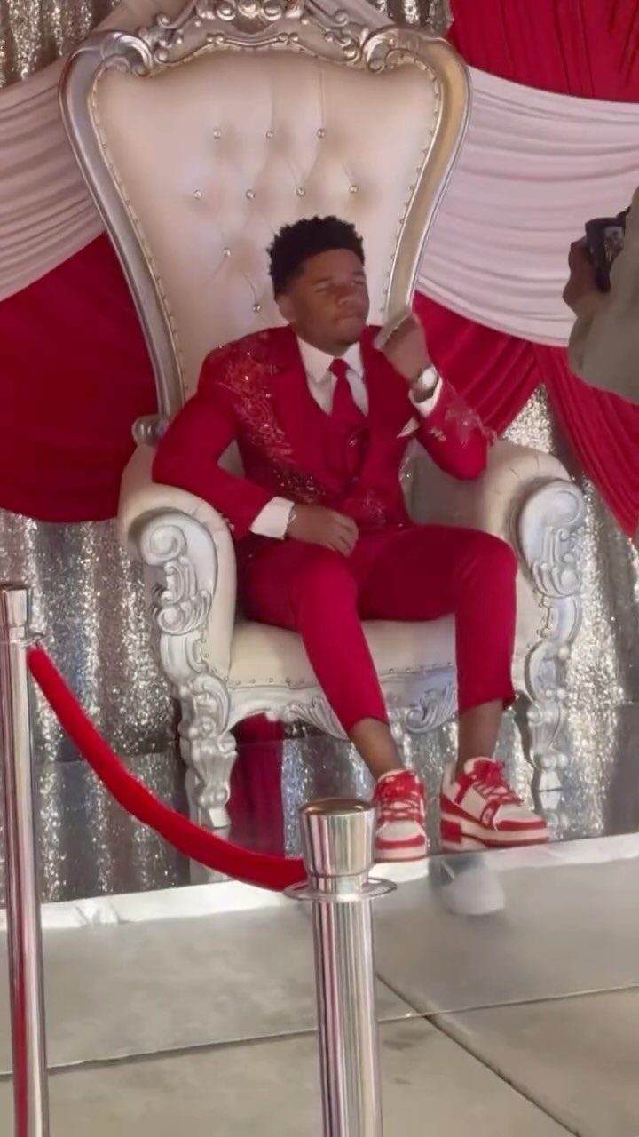 Young man in an embroidered red suit sitting on a white ornate throne.