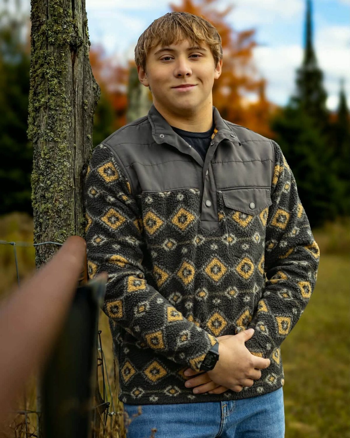 Young man in patterned fleece leaning against a tree in autumn landscape