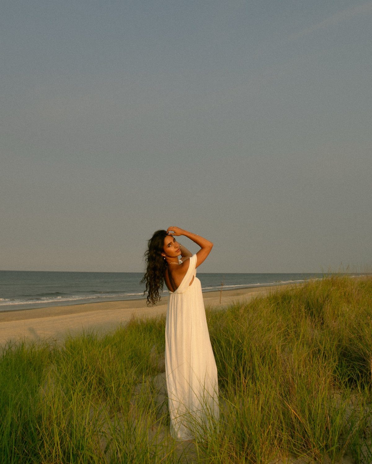 Woman in a white dress posing in beach grass by the ocean shore.