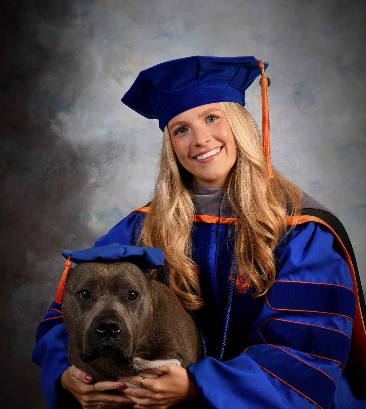 Graduating woman in blue gown holding a grey dog wearing a graduation cap.