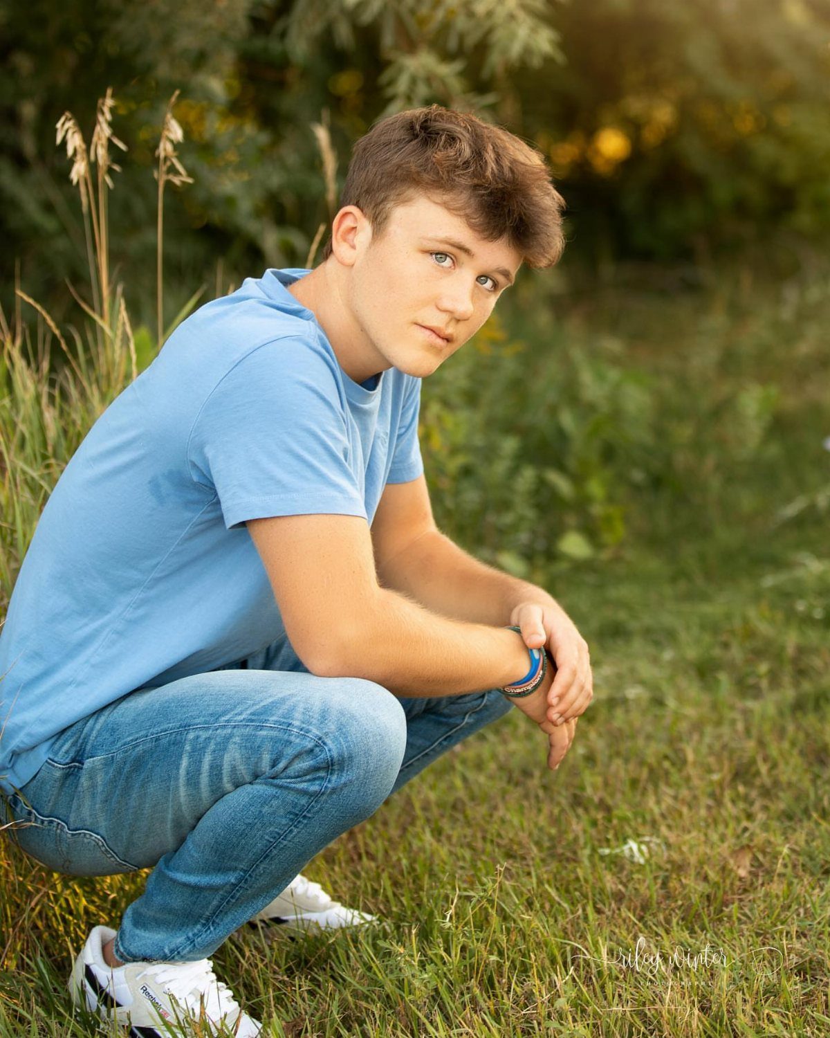 Teenage boy in blue shirt and jeans squatting in a sunny grassy field