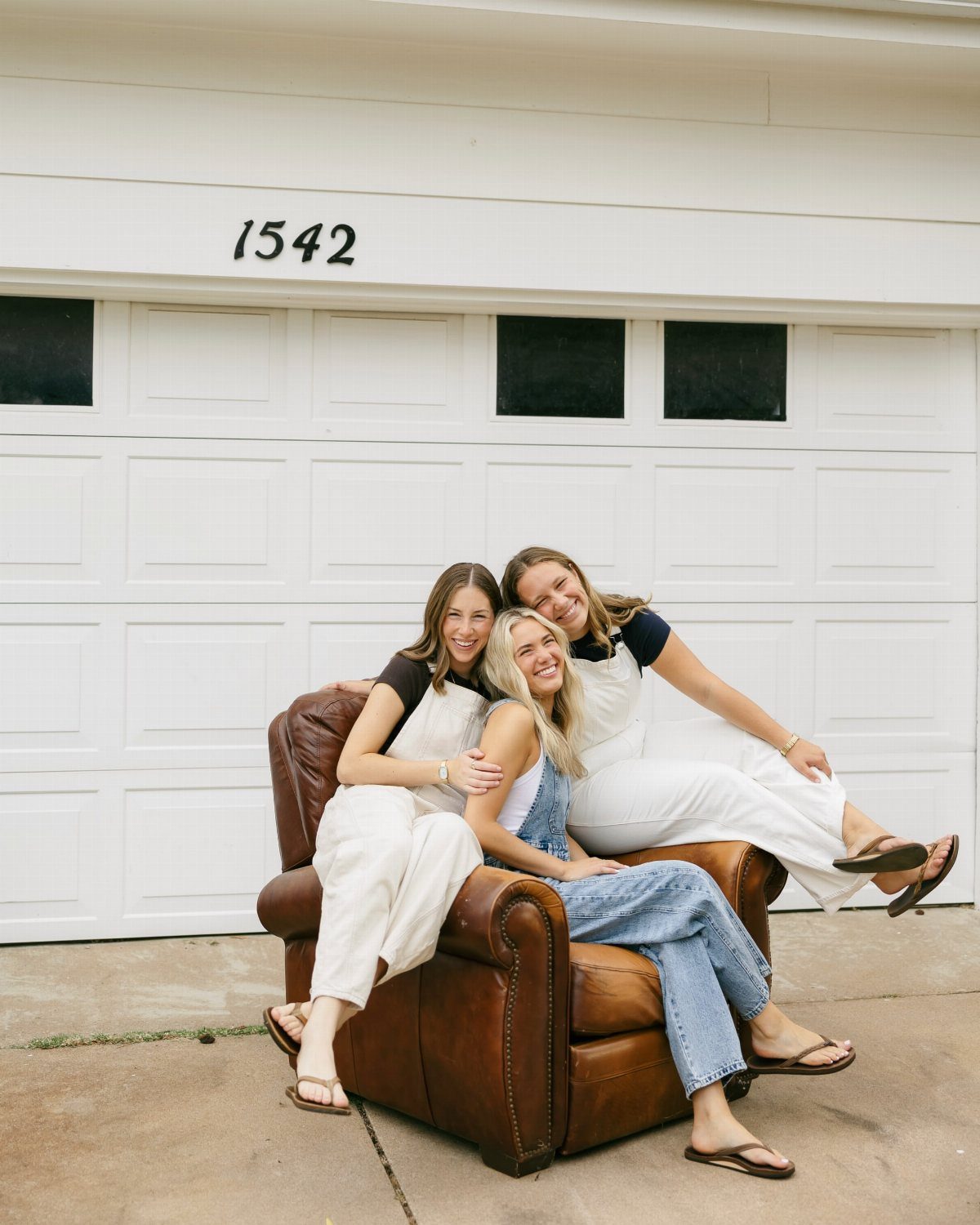 Three smiling young women posing together in a leather armchair outdoors.