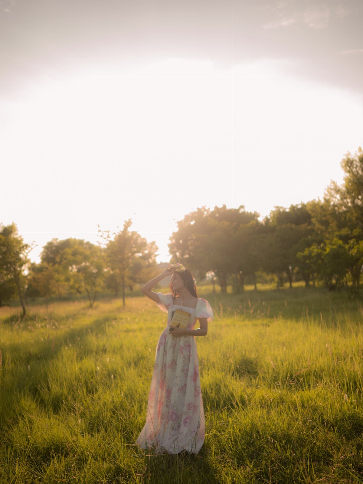 Young woman in a white floral dress standing in a sunlit grassy field.