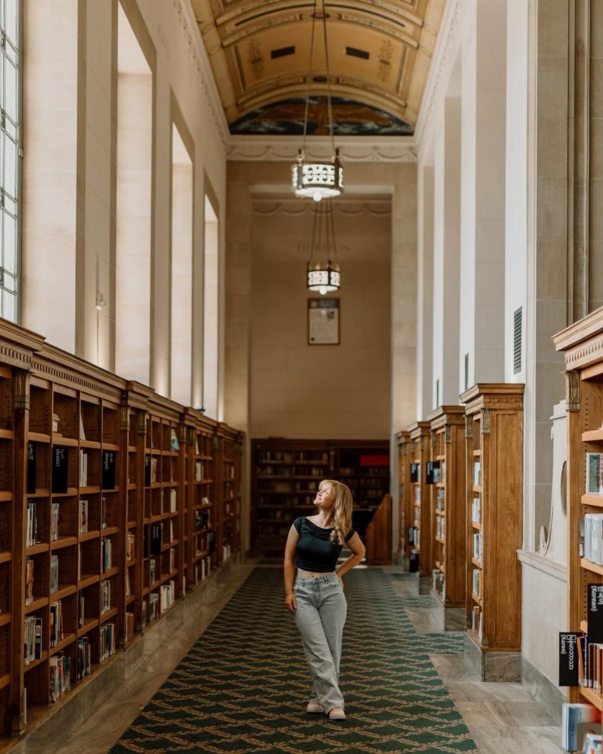 Young woman posing in a grand library with tall wooden bookshelves and ornate ceilings.