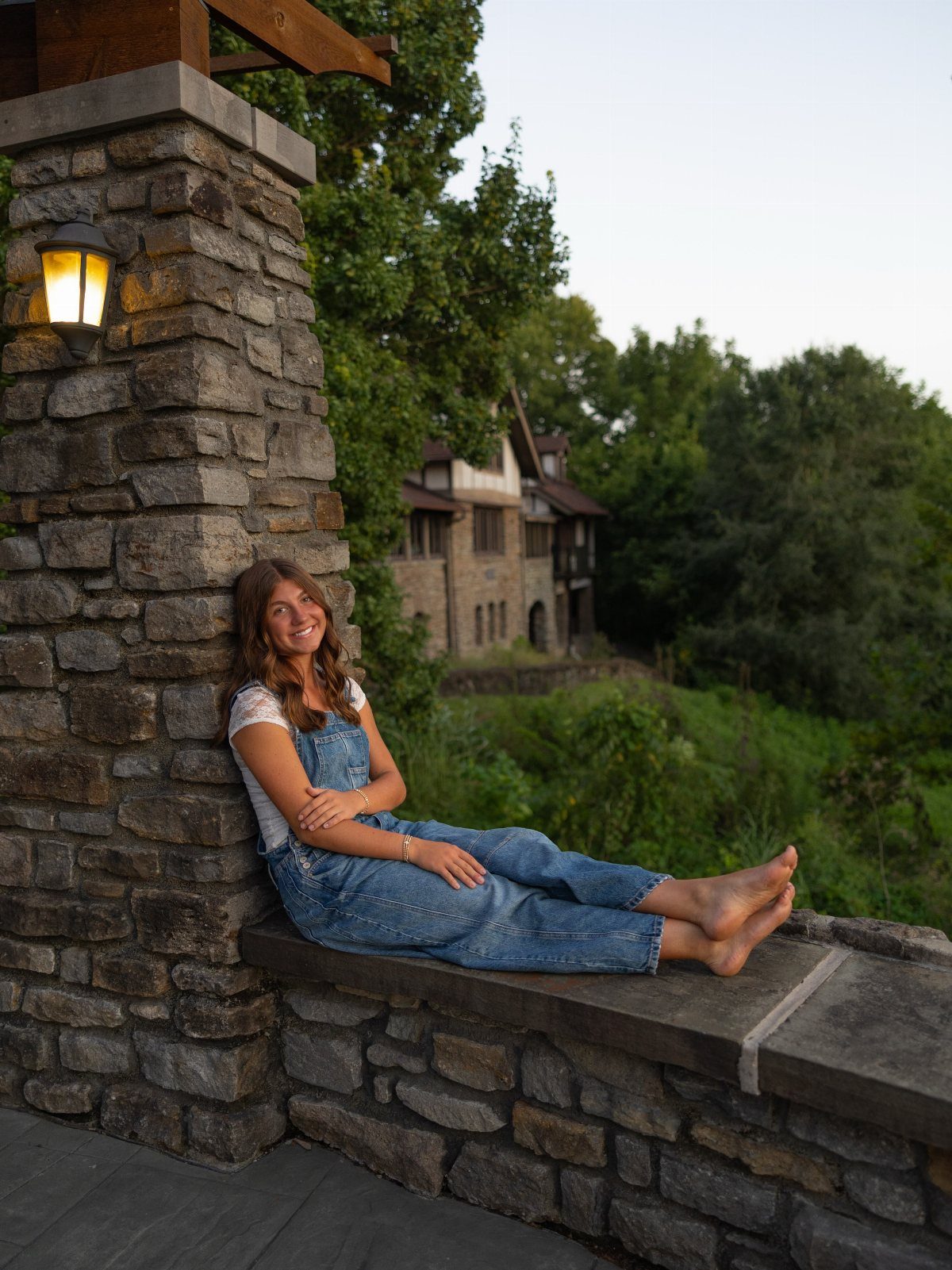 Young woman in denim overalls sitting on a stone ledge near a building.