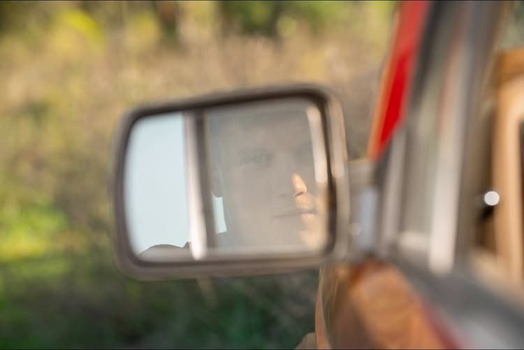 Person's face reflected in a car side-view mirror with blurred nature background