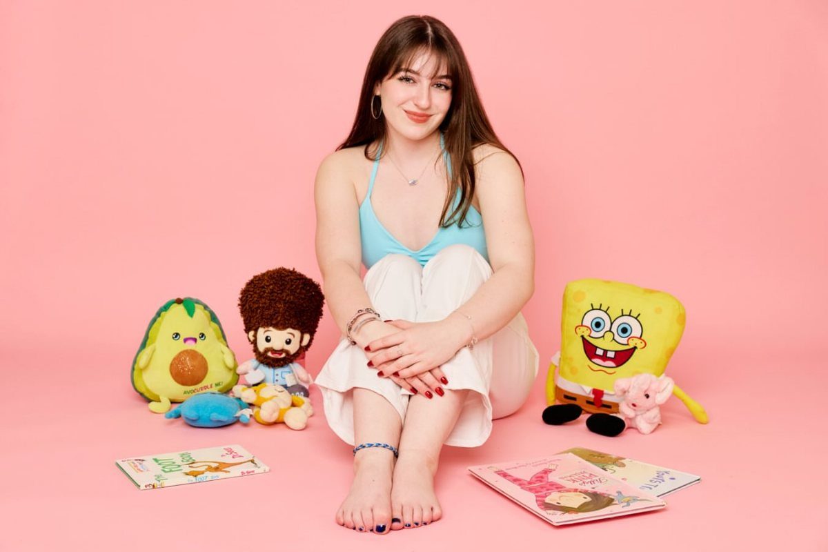 Young woman sitting on a pink background surrounded by plush toys and books.