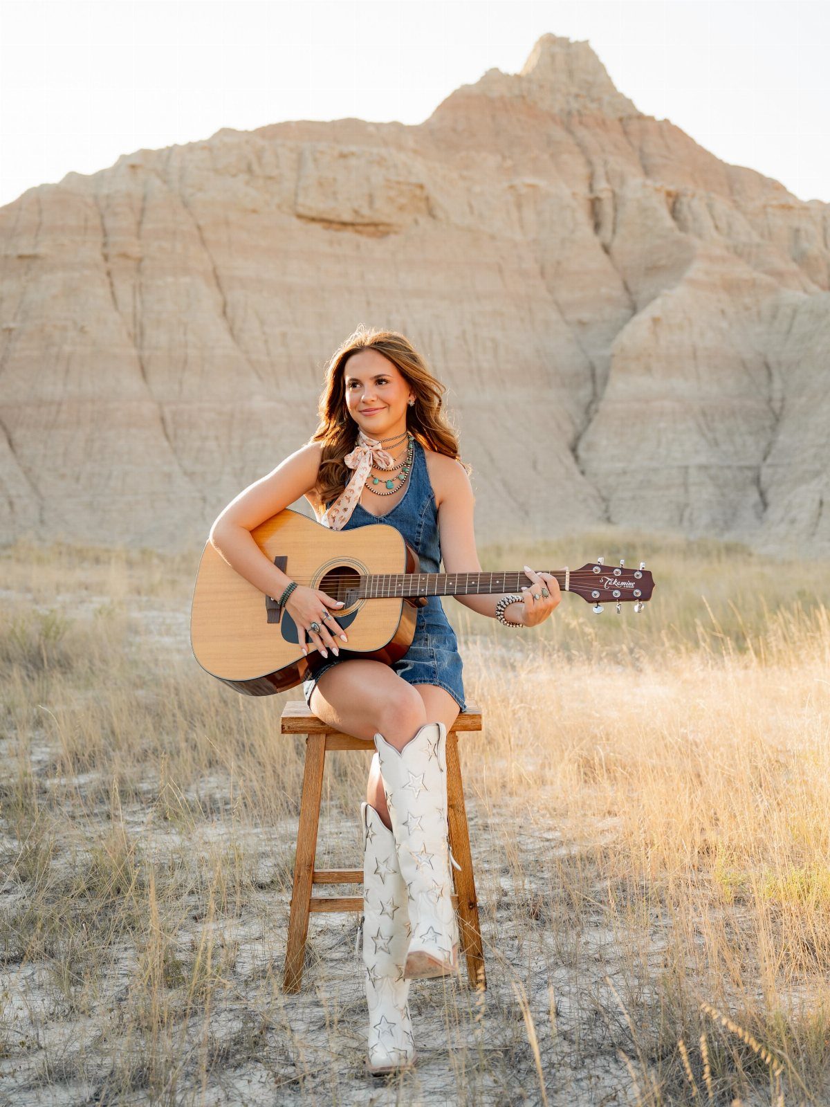 Young woman in a denim dress and white boots playing guitar in desert