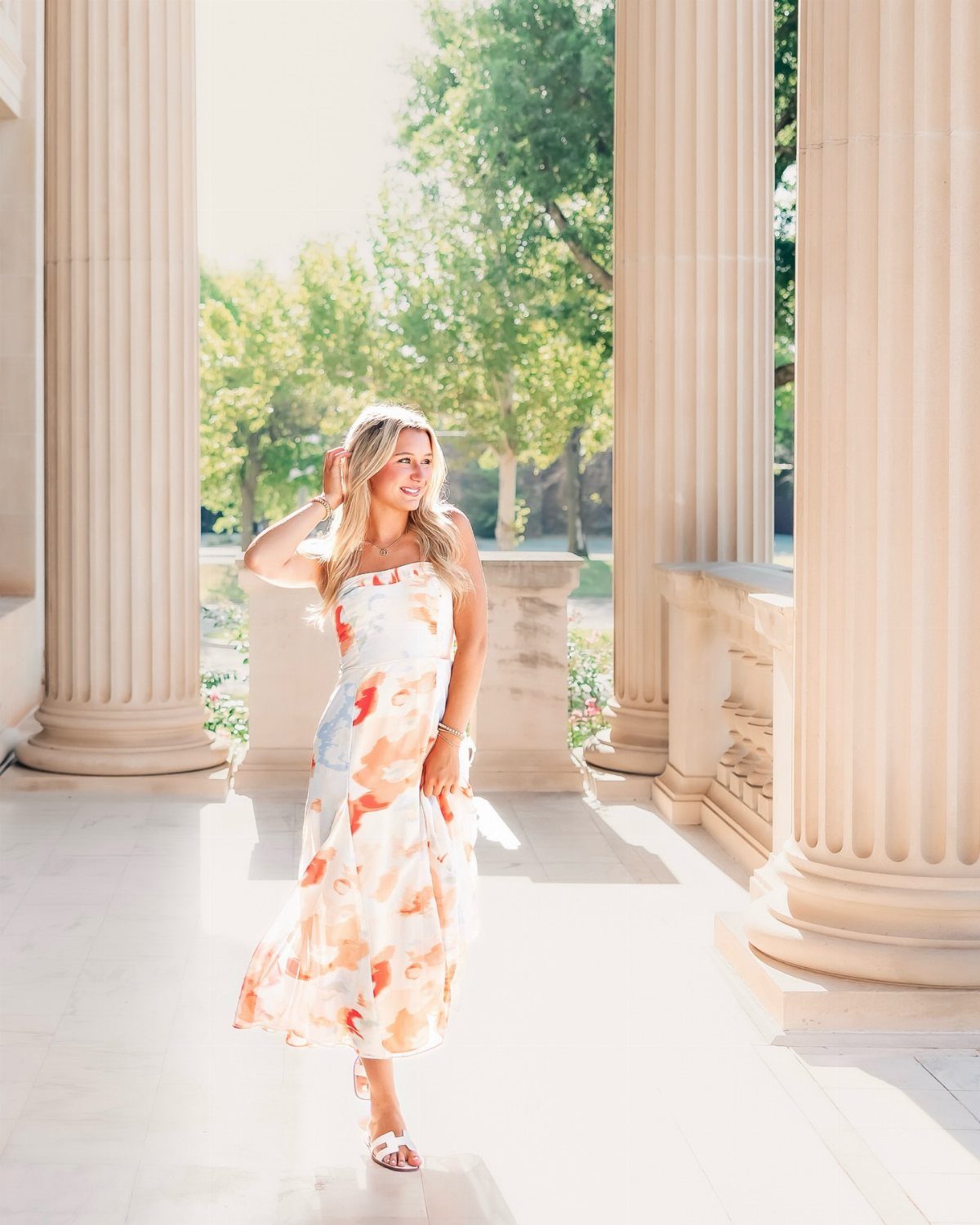 Young woman in a long flowy watercolor dress posing between stone columns.