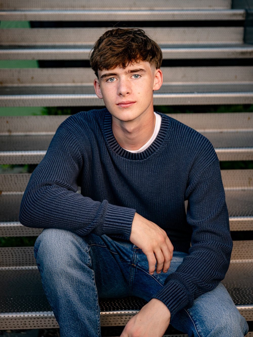 Young man in a blue knit sweater and jeans sitting on metal stairs.