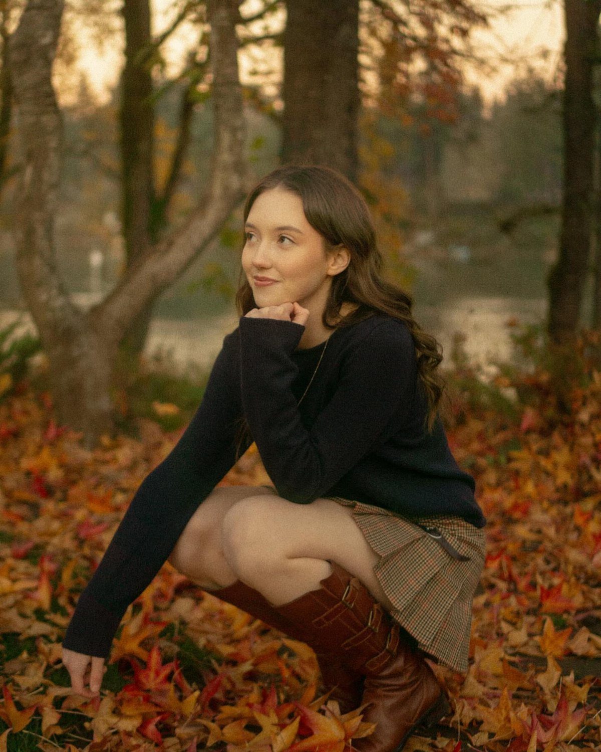 Young woman in navy sweater and plaid skirt sitting in autumn leaves.