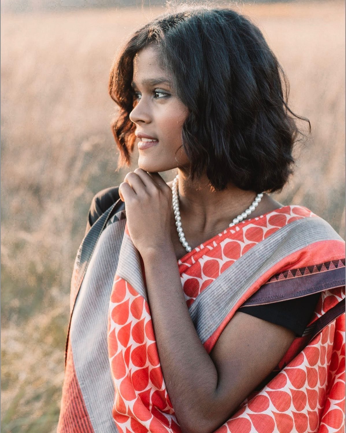 Young woman in a red patterned saree and pearls posing in a field.