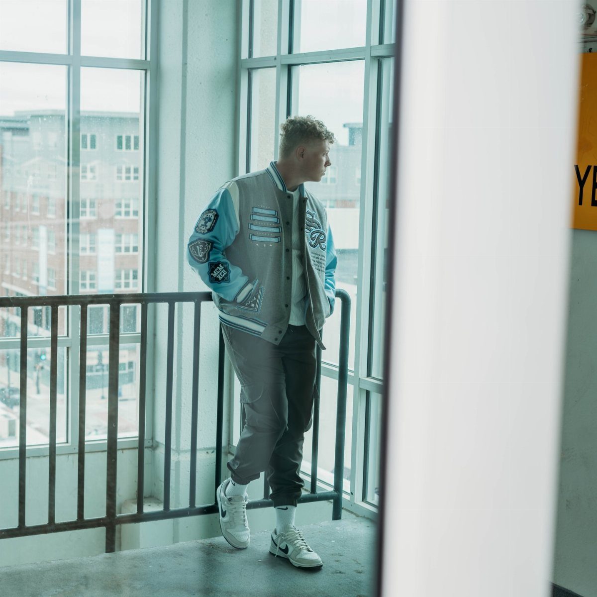 Young man wearing a blue varsity jacket and grey trousers by a window