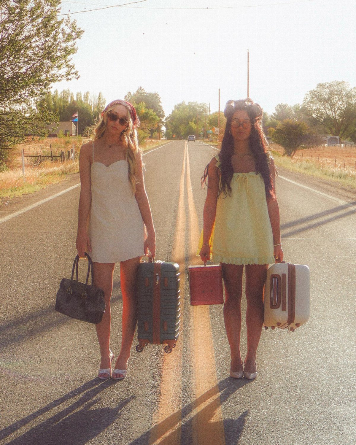 Two women in short dresses holding vintage suitcases on a rural road.