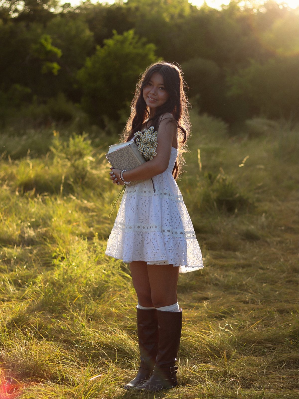 Young woman in white eyelet dress and brown boots posing in sunny field