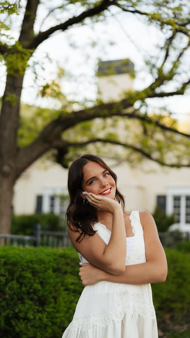 Young woman in a white dress posing and smiling outdoors near a tree