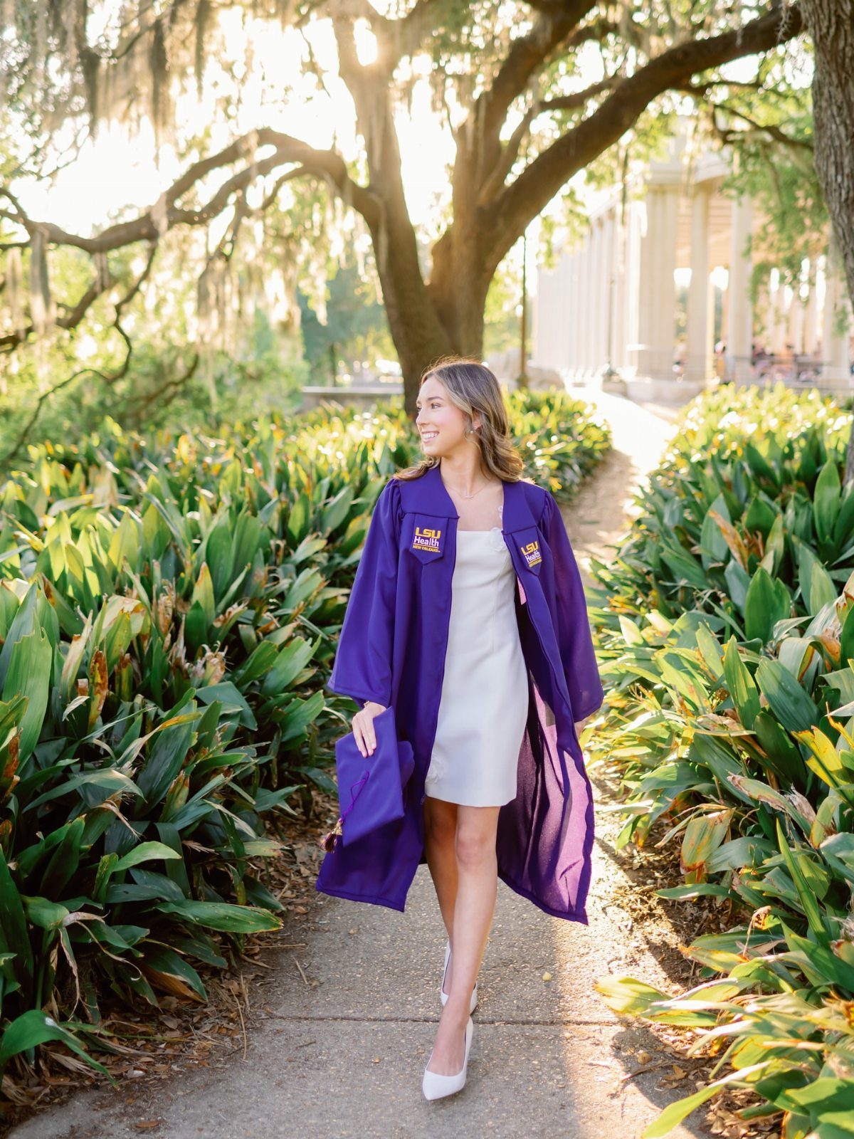 Young woman in purple graduation gown walking along a sunny garden path