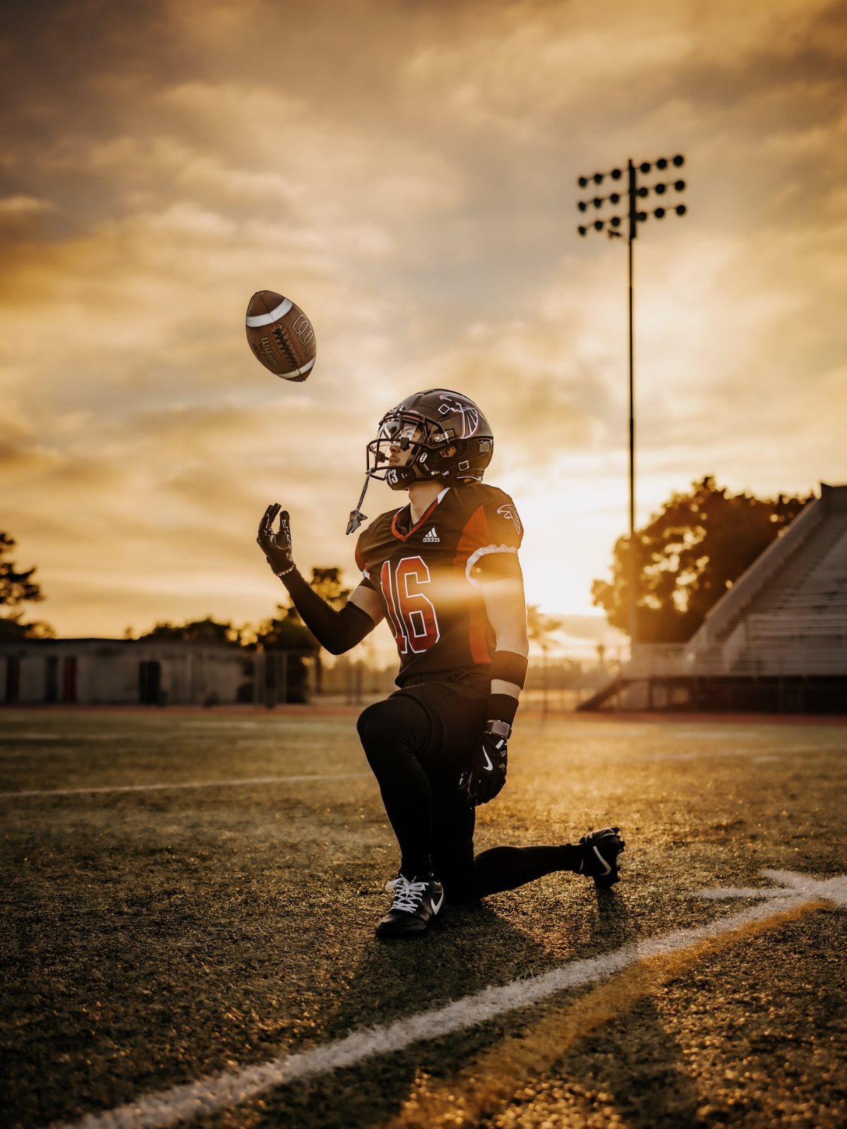 Football player kneeling on a field looking up at a flying football at sunset