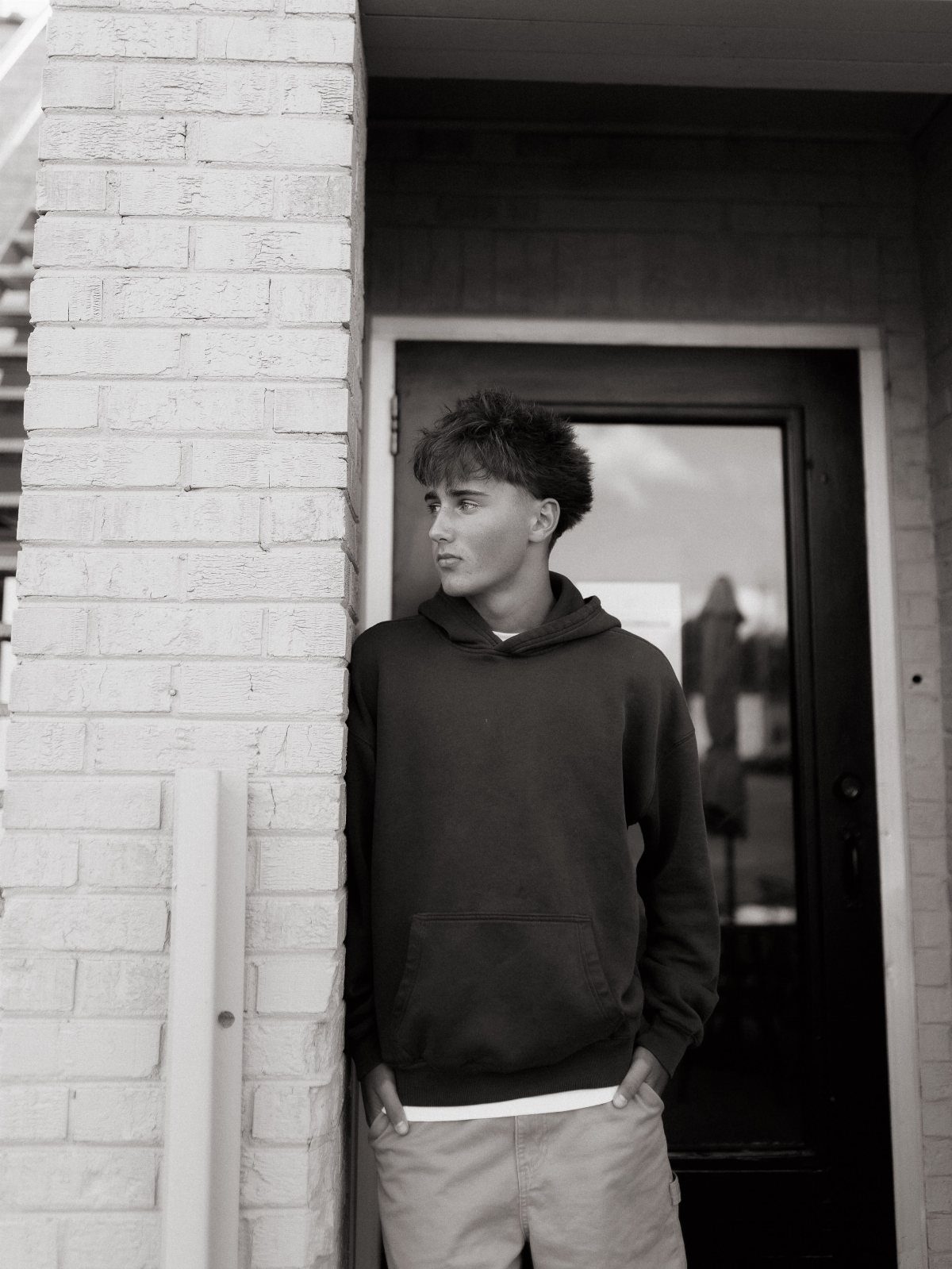 Young man in a hoodie leaning against a white brick wall in monochrome.
