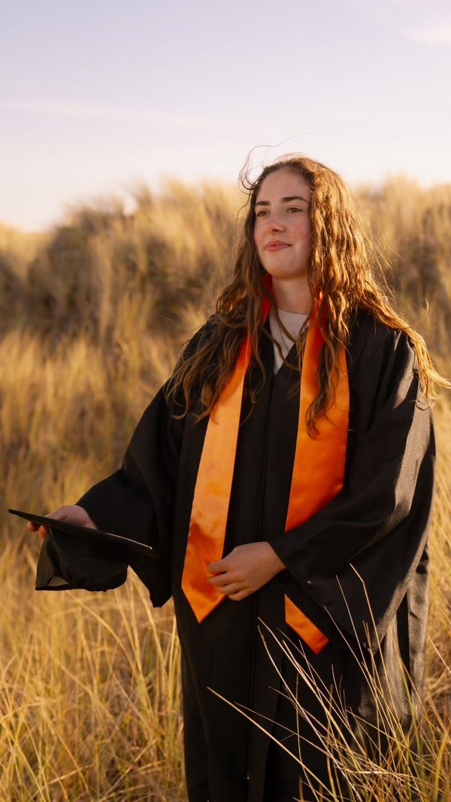 Young woman in black graduation gown and orange stole standing in golden grass